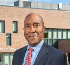 Smiling man in a suit stands in front of a modern brick building with large windows.