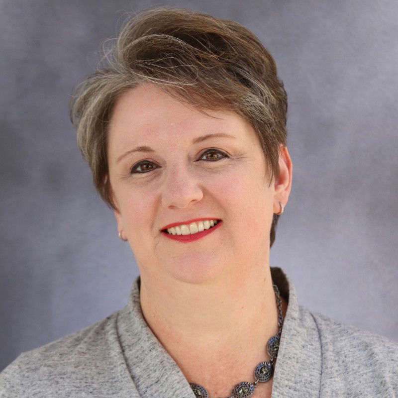 Smiling woman with short gray hair, wearing a gray sweater and necklace against a neutral background.