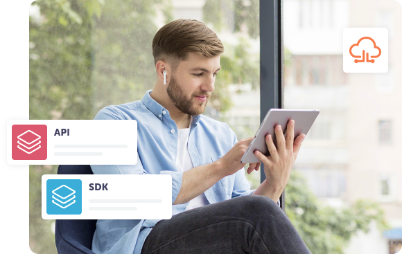 Vīrietis ar planšetdatoru sēž pie liela loga, ausīs ieliktas austiņas. Pārklājumā redzamas ikonas ar uzrakstiem “API” un “SDK”, kas izceļ programmatūras integrāciju, savukārt mākoņa ikona simbolizē mākoņdatošanas ieviešanu.