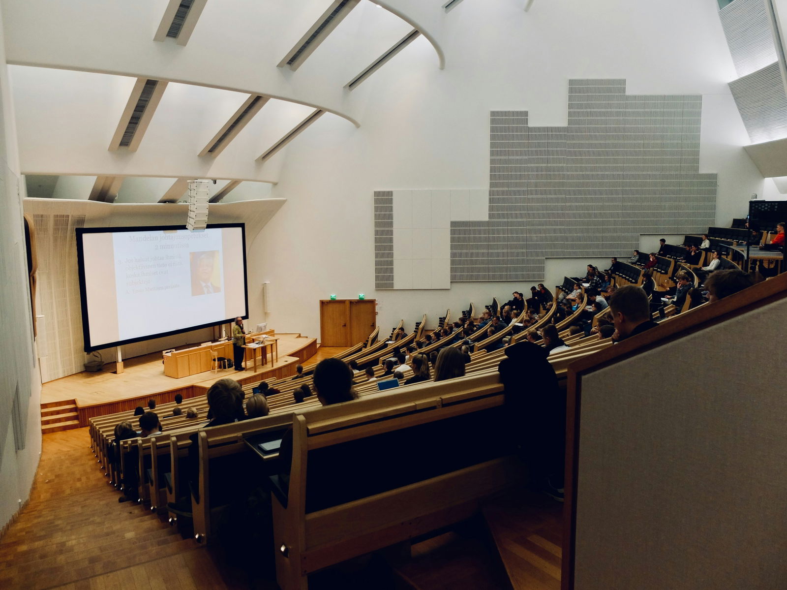 Lecture hall with audience, presenter at podium, and large screen displaying slides.