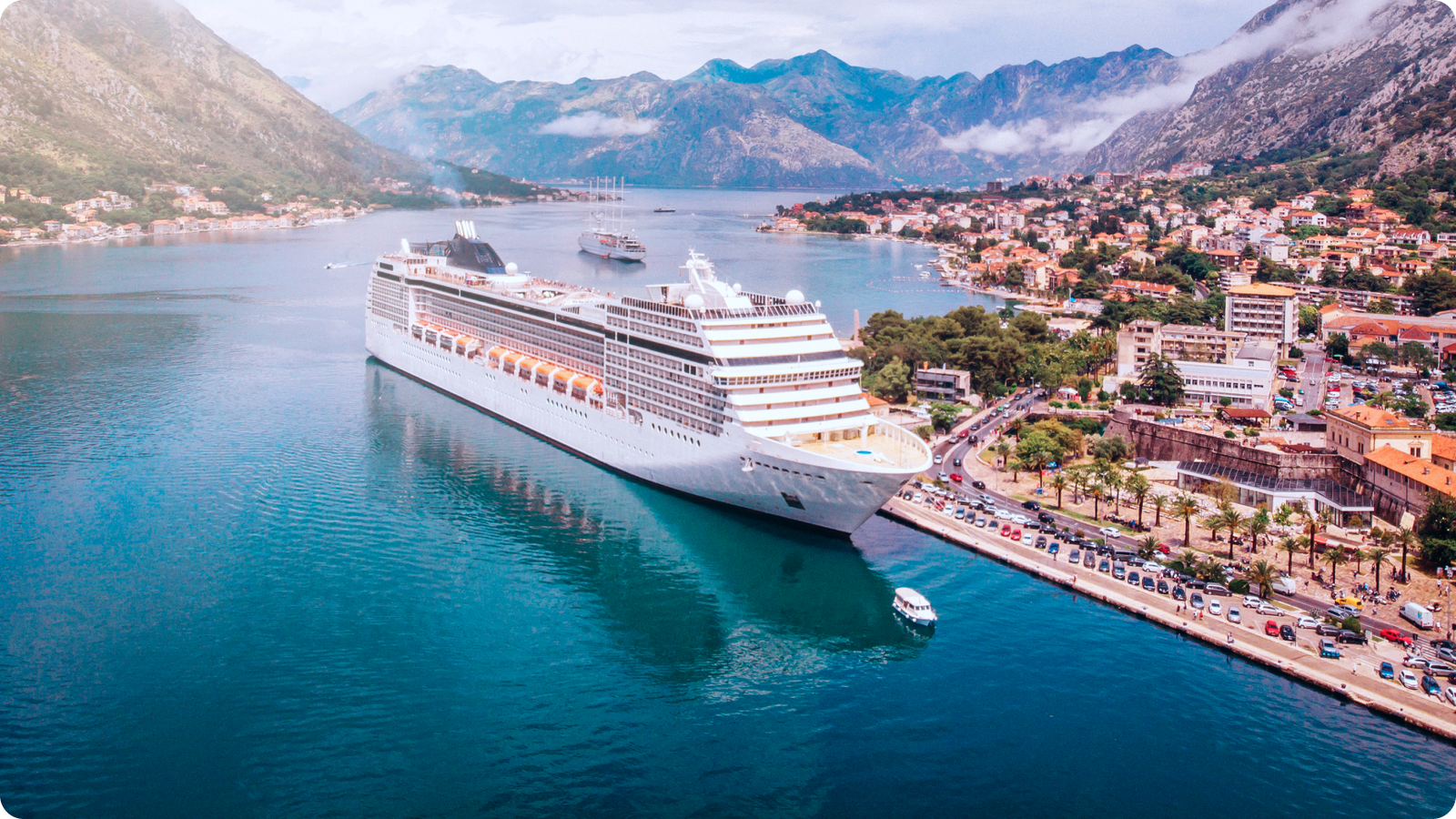 Aerial view of a large cruise ship docked near a scenic coastal town and mountains.