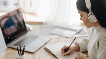 Persoon met een laptop open voor zich en koptelefoons op maakt aantekeningen in een schrift.