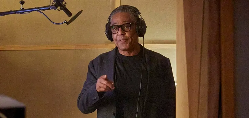 L'attore Giancarlo Esposito, noto per i ruoli in Breaking Bad e The Mandalorian, è in piedi in uno studio di registrazione e indossa le cuffie; è rivolto verso l'ascoltatore. Alla sua sinistra è visibile un microfono.