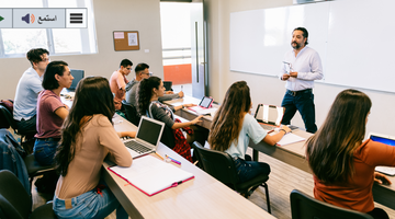 Students in a classroom listen to the teacher standing at the board.