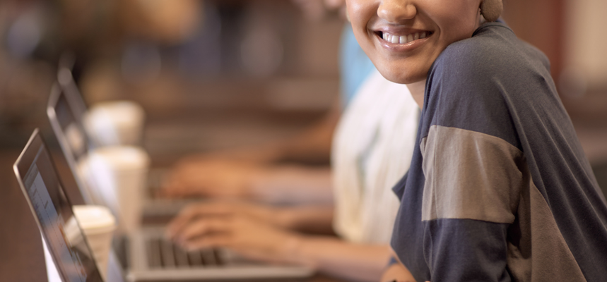 Smiling learner sitting at a laptop, with others typing on computers in the background.