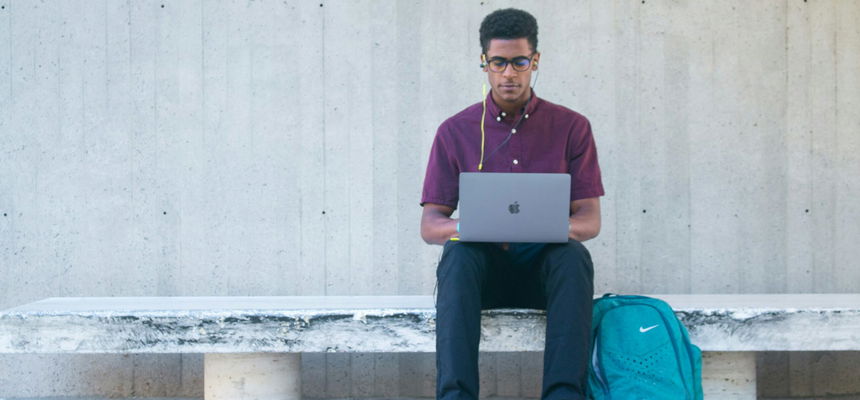 A man sitting on a bench with a laptop and blue backpack, wearing glasses and a purple shirt.