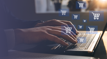 A person is typing on their laptop with shopping cart icons floating above the keyboard.