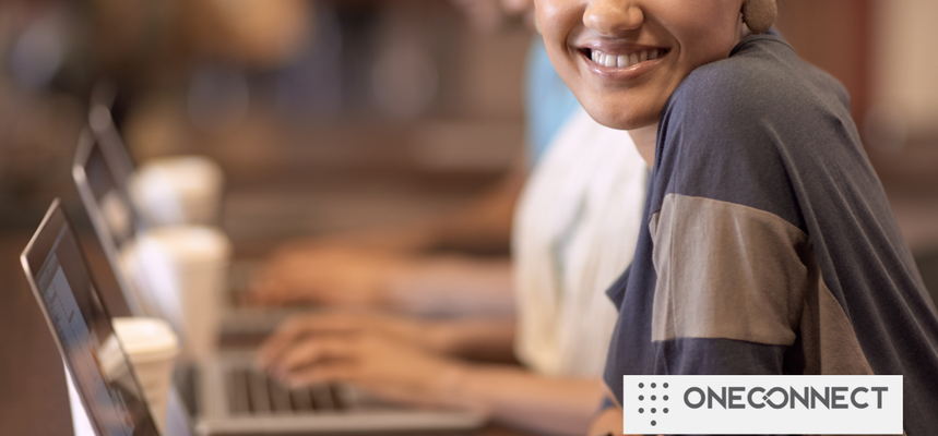 A woman smiles at the camera while typing on a laptop in a cafe. Warm ambient lighting and blurred background suggest a relaxed atmosphere. The text "ONECONNECT" is in the corner.