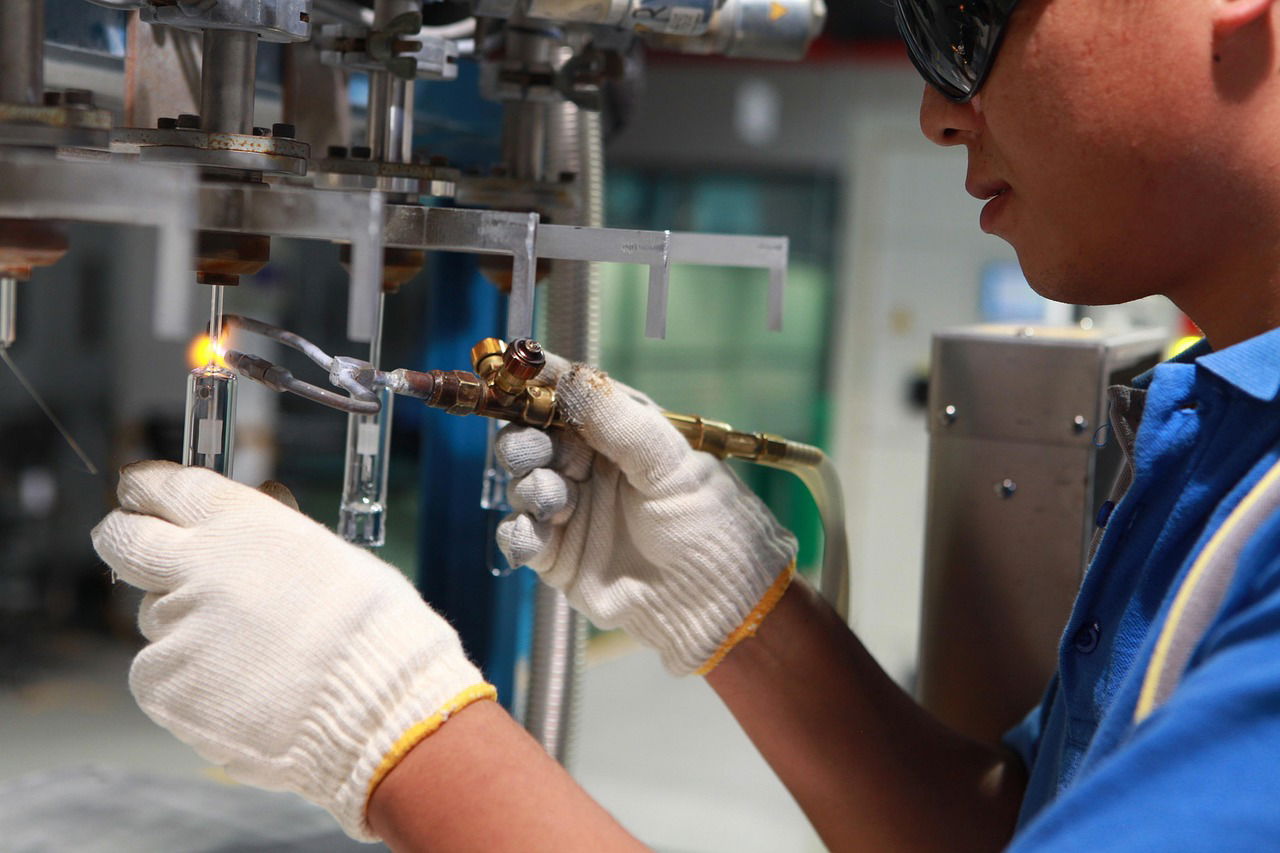 Worker wearing safety gloves and goggles using a gas torch to weld glass components in an industrial setting.