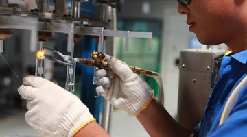 Worker wearing safety gloves and goggles using a gas torch to weld glass components in an industrial setting.