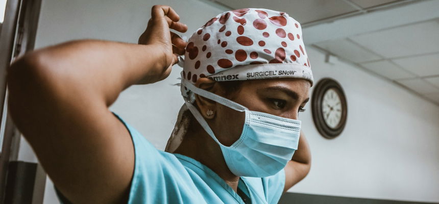 Woman wearing blue surgical scrubs and cap, tying a white face mask behind her head.