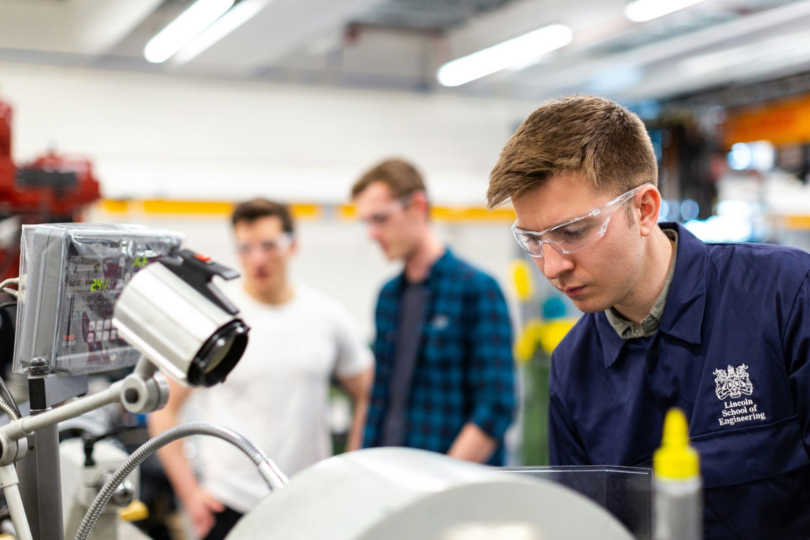 Engineering students, wearing goggles, standing by machinery, representing accessibility in vocational training.