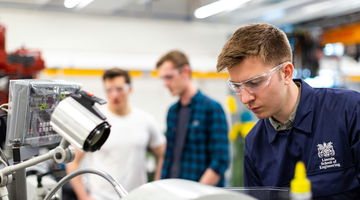 Engineering students, wearing goggles, standing by machinery, representing accessibility in vocational training.