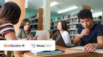 Alunos sentados à mesa em uma biblioteca, aprendendo com livros, laptops e celulares.