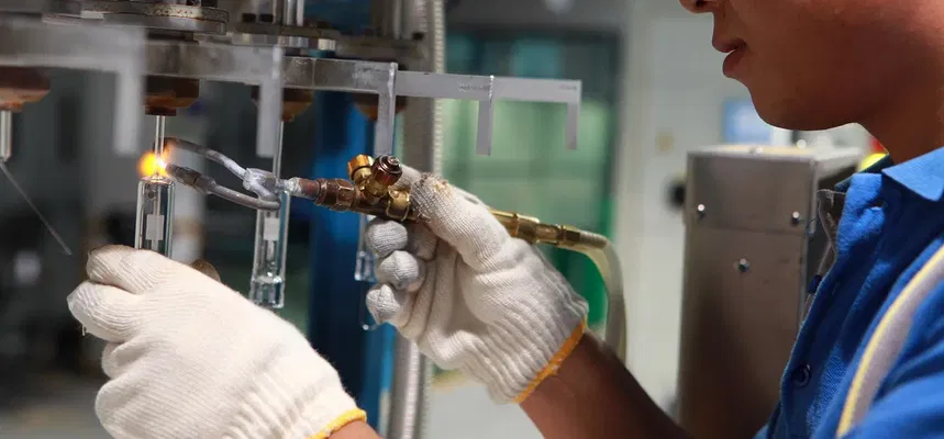 Un travailleur avec des gants et des lunettes de sécurité utilisant une torche à souder pour assembler des éléments en verre dans un atelier industriel.