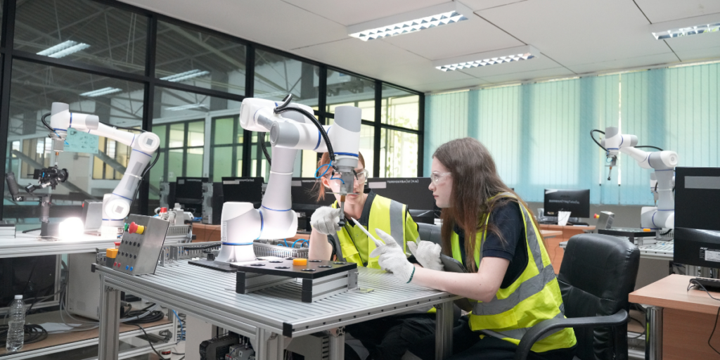 Two young women wearing hi-visibility jackets and googles sitting at a desk in a high-tech engineering lab, with robotic machinery on the desks around them. 
