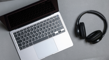 A photo of a silver laptop and black headphones on a grey desk.