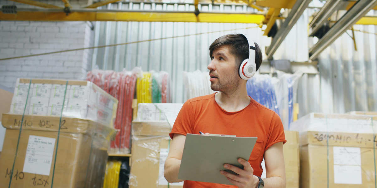 A young man in a clothing warehouse, with a rack of clothes behind him. He is dressed in an orange t-shirt, and is wearing headphones and holding a clipboard.
