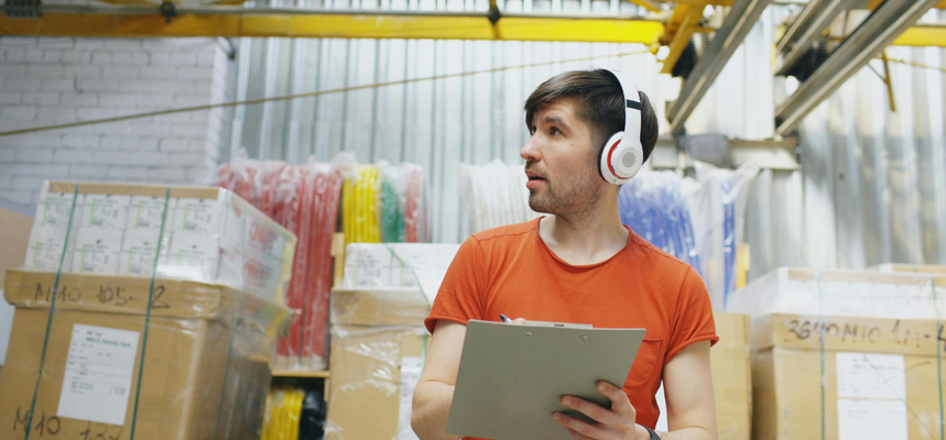 A young man in a clothing warehouse, with a rack of clothes behind him. He is dressed in an orange t-shirt, and is wearing headphones and holding a clipboard.
