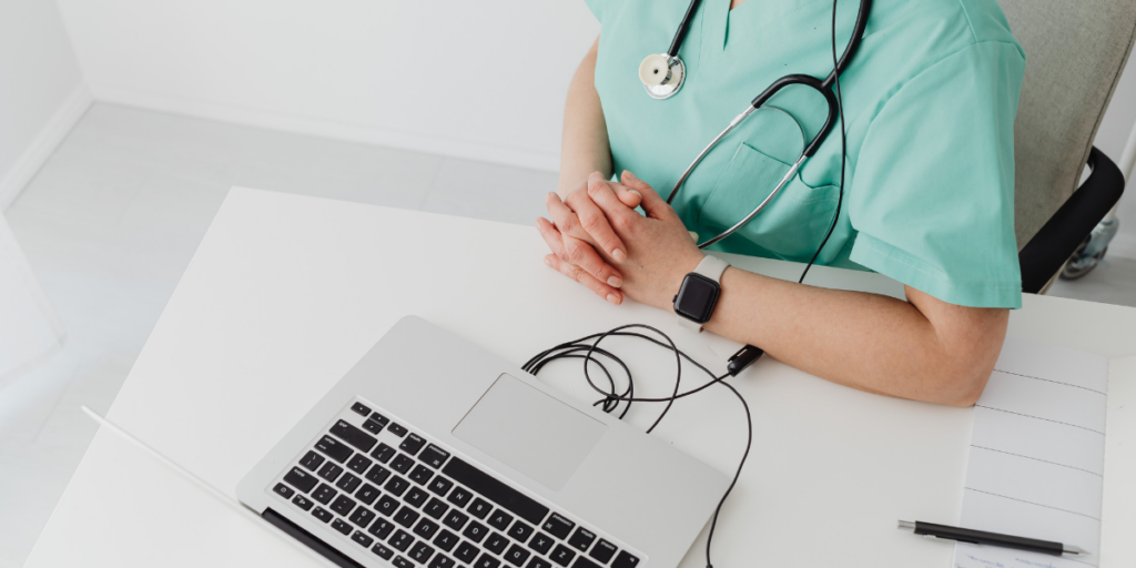 Medical student or professional wearing pale green scrubs, with a stethoscope around her neck, and the wire from headphones connecting to a laptop sitting on a desk in front of her. 