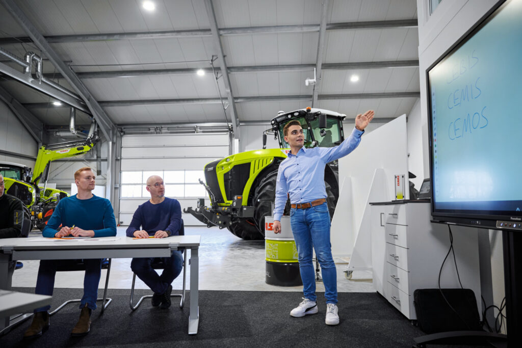 A man explaining concepts on a screen to other two men. There are two bright green tractors behind them, inside of CLAAS' facilities.