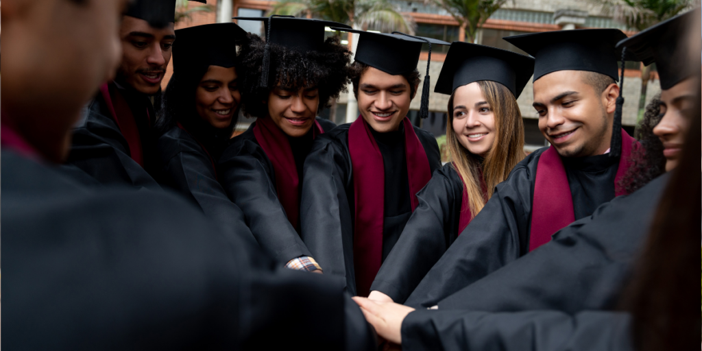 A group of 8 or 9 higher education students standing in a semi-circle, wearing black mortar boards and gowns, and celebrating graduation.