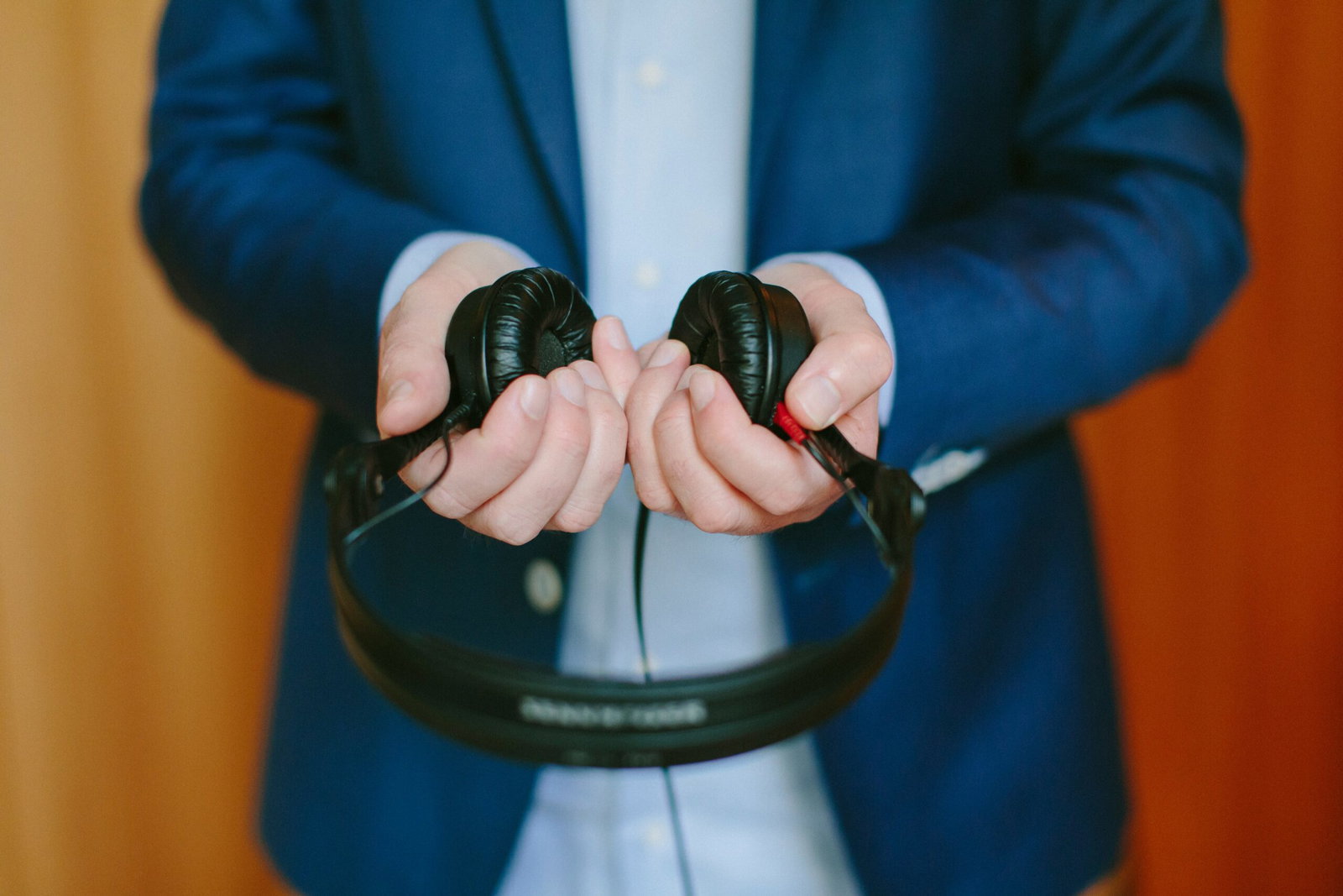 A man wearing a dark blue business suit and pale blue shirt, holding black headphones our in front of him.