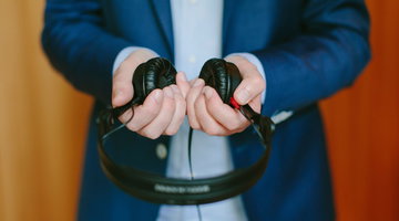 A man wearing a dark blue business suit and pale blue shirt, holding black headphones our in front of him.
