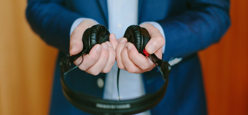 A man wearing a dark blue business suit and pale blue shirt, holding black headphones our in front of him.