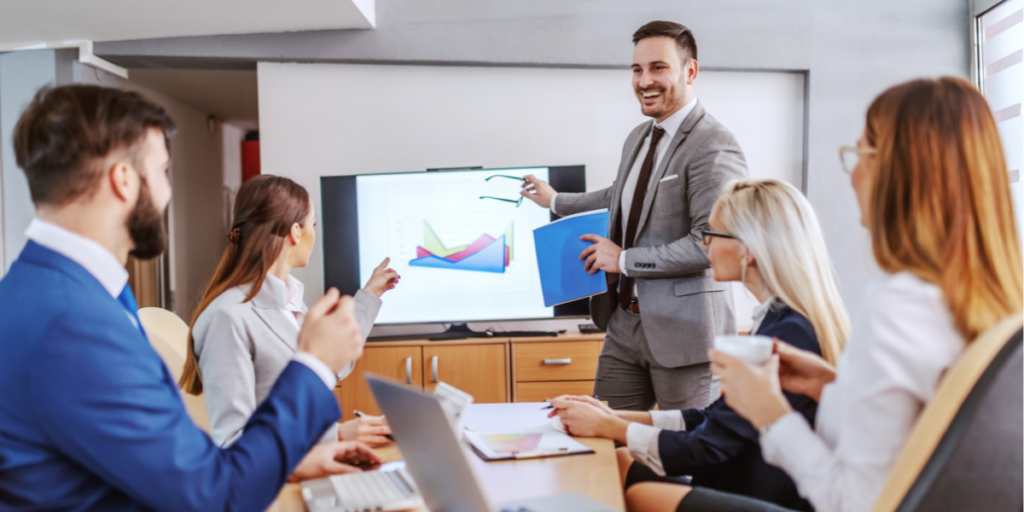 A corporate boardroom, with four employees sitting at a boardroom table with lapts, and a fifth smiling man in a grey suit standing making a presentation to them. in front of a large screen.