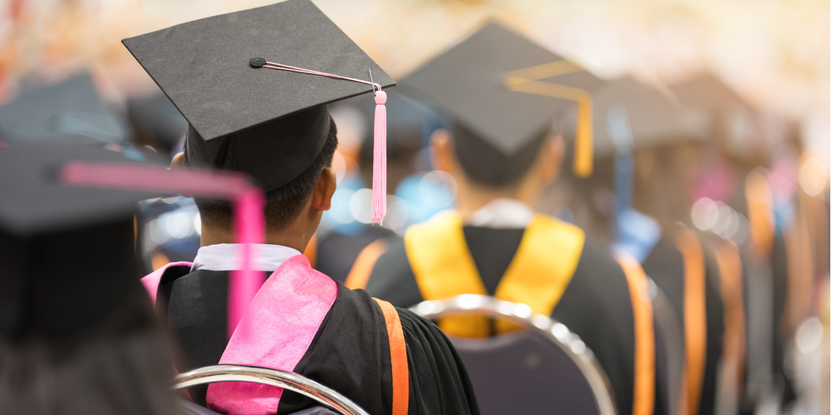 University students on graduation day, sitting in rows with their backs to the camera, wearing mortar boards and gowns.