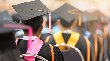 University students on graduation day, sitting in rows with their backs to the camera, wearing mortar boards and gowns.
