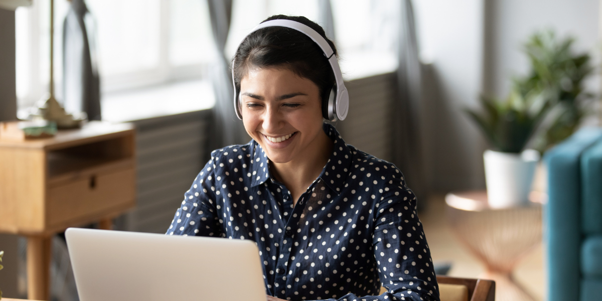 A smiling and smartly-dressed woman wearing a navy and white spotted blouse, sitting at a desk with a laptop and wearing headphones.
