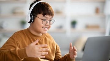 A boy of around 12 or 13 sits looking and smiling at a laptop. He is wearing headphones and his hands are gesturing towards the screen.
