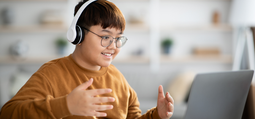 A boy of around 12 or 13 sits looking and smiling at a laptop. He is wearing headphones and his hands are gesturing towards the screen.