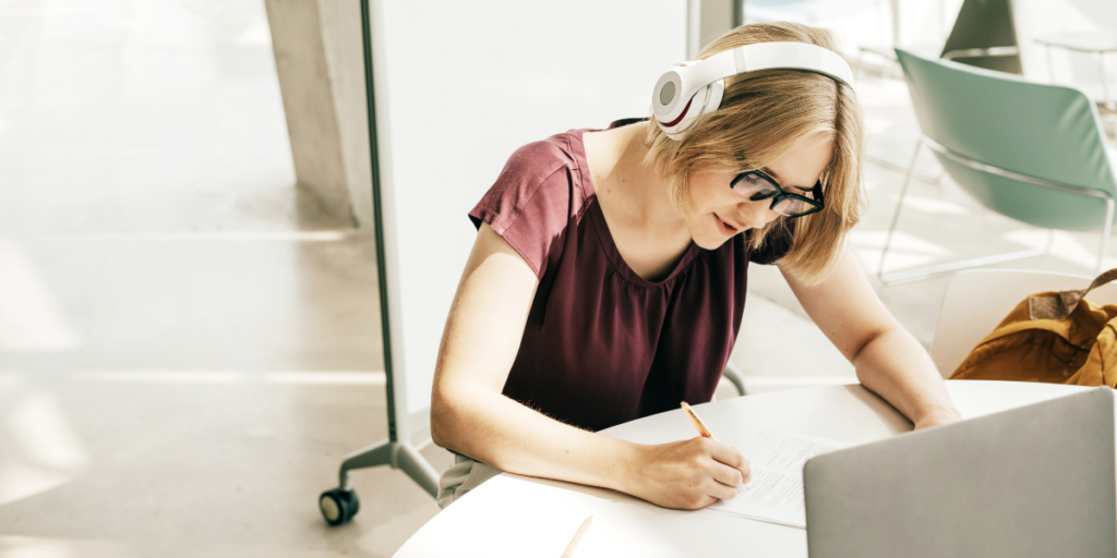 A young woman sits at a white table writing notes, in front of a laptop, while wearing white headphones.