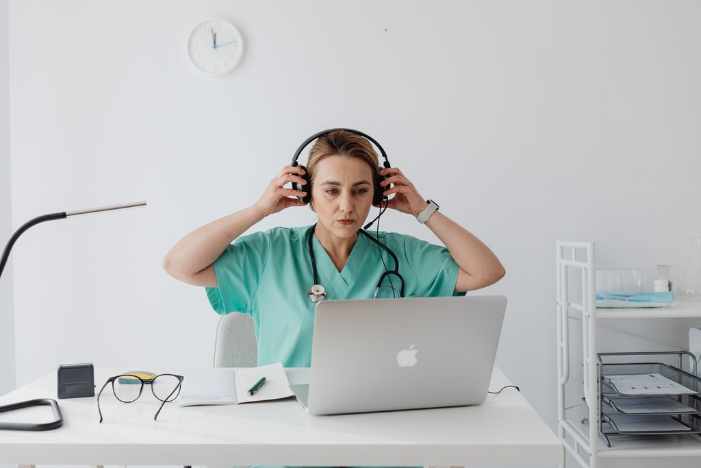 A healthcare worker in scrubs sits at a desk, putting on headphones while looking at a laptop. A notepad, glasses, and pen are on the desk.