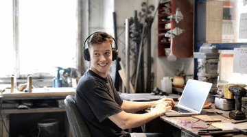 A young man wearing headphones smiles while working on a laptop at a cluttered workshop desk with tools and materials around him.