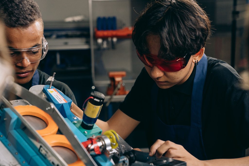Two young students wearing safety goggles and aprons operate a piece of machinery with orange wheels in a workshop or industrial setting.