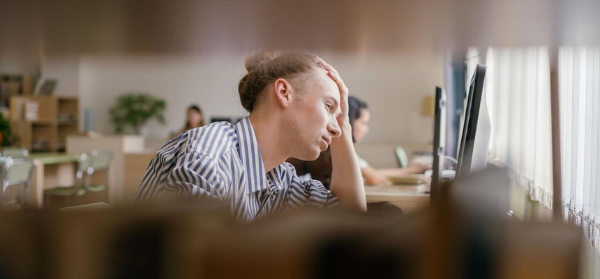 A man seated at a desk, focused on his computer screen, with a notepad and pen nearby. He looks focused, and his surroundings are blurry.