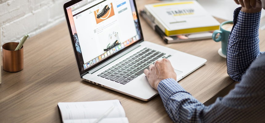 A man focused on his laptop, seated at a desk with a notepad and some books nearby.