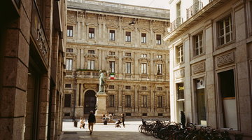 Una via stretta di Milano che si apre su una piccola piazza, dove una statua di bronzo su un basamento di pietra si trova davanti a un palazzo signorile con la bandiera italiana. Alcuni passanti attraversano la pavimentazione e una lunga fila di biciclette e parcheggiata sul lato destro.