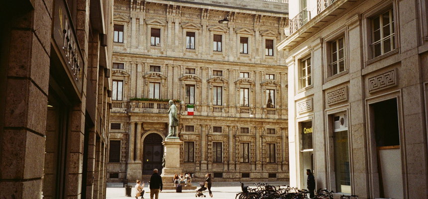 Una via stretta di Milano che si apre su una piccola piazza, dove una statua di bronzo su un basamento di pietra si trova davanti a un palazzo signorile con la bandiera italiana. Alcuni passanti attraversano la pavimentazione e una lunga fila di biciclette e parcheggiata sul lato destro.