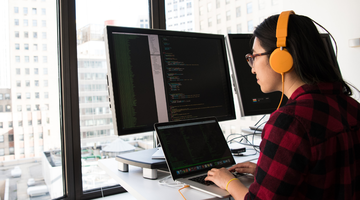 A developer wearing bright orange headphones works at a standing desk beside a city window, typing on a laptop in front of two large monitors filled with source code.