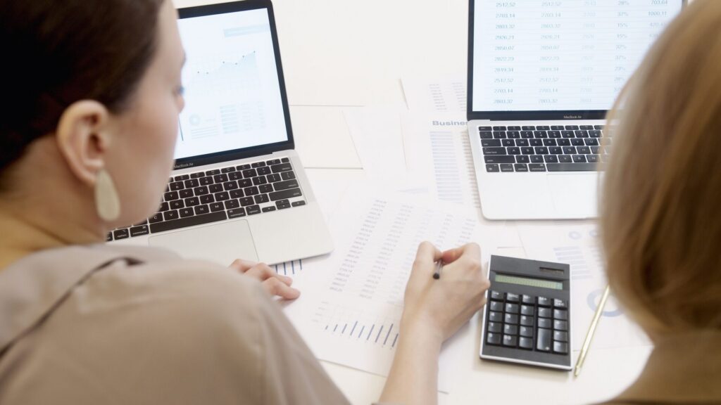 Two colleagues seen from behind reviewing charts and spreadsheets on two laptops, with printed data sheets and a calculator spread across the desk.