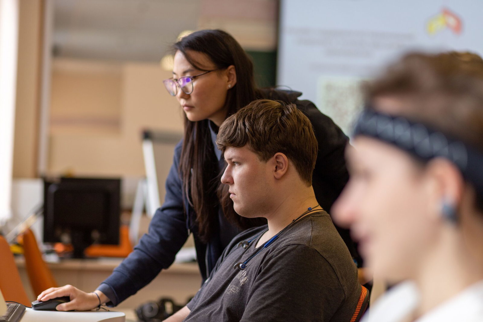 A teacher leans over a desk to help a student working at a computer, with two more students at screens beside them, headphones around their necks.