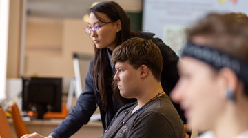 A teacher leans over a desk to help a student working at a computer, with two more students at screens beside them, headphones around their necks.