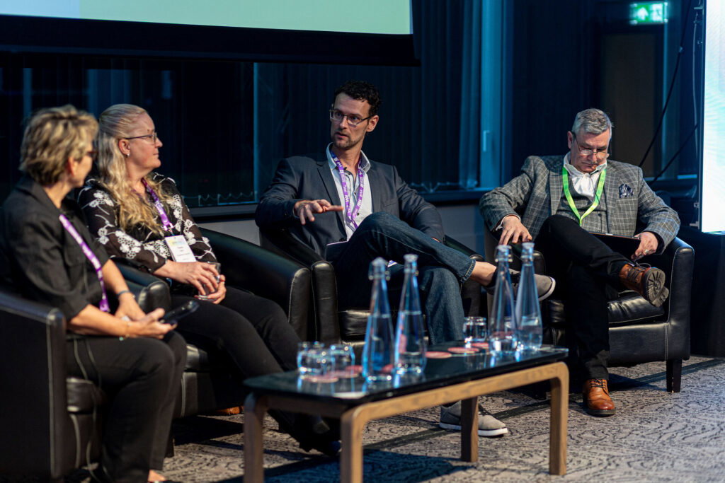 Four panellists seated in armchairs on a conference stage, one speaking and gesturing with his hand, conference lanyards around their necks.